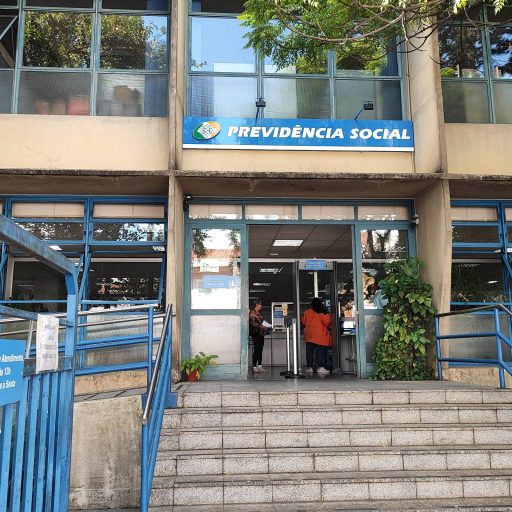 m mostra a entrada de uma agência da Previdência Social. A fachada é de um prédio de dois andares, com janelas grandes e uma escada de acesso. Acima da entrada,