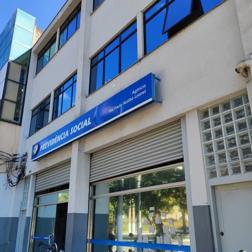 m mostra a fachada de um prédio de três andares. Na parte superior, há janelas grandes e um letreiro azul com a inscrição 'PREVIDÊNCIA SOCIAL'. Abaixo do letrei